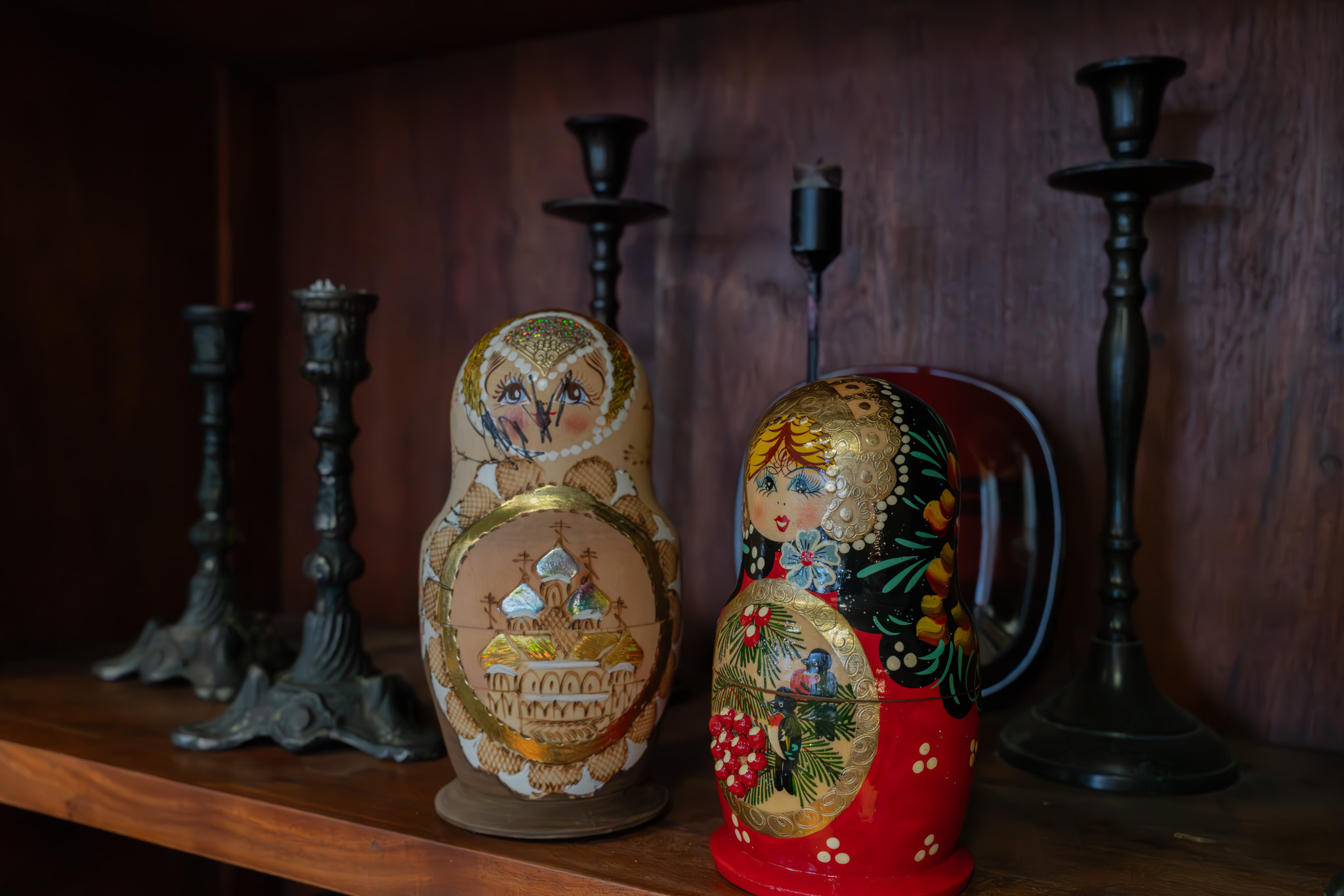 Kaksi koristeltua venäläistä matryoshka-nukkea puisella hyllyllä, taustalla useita tummia kynttilänjalkoja.