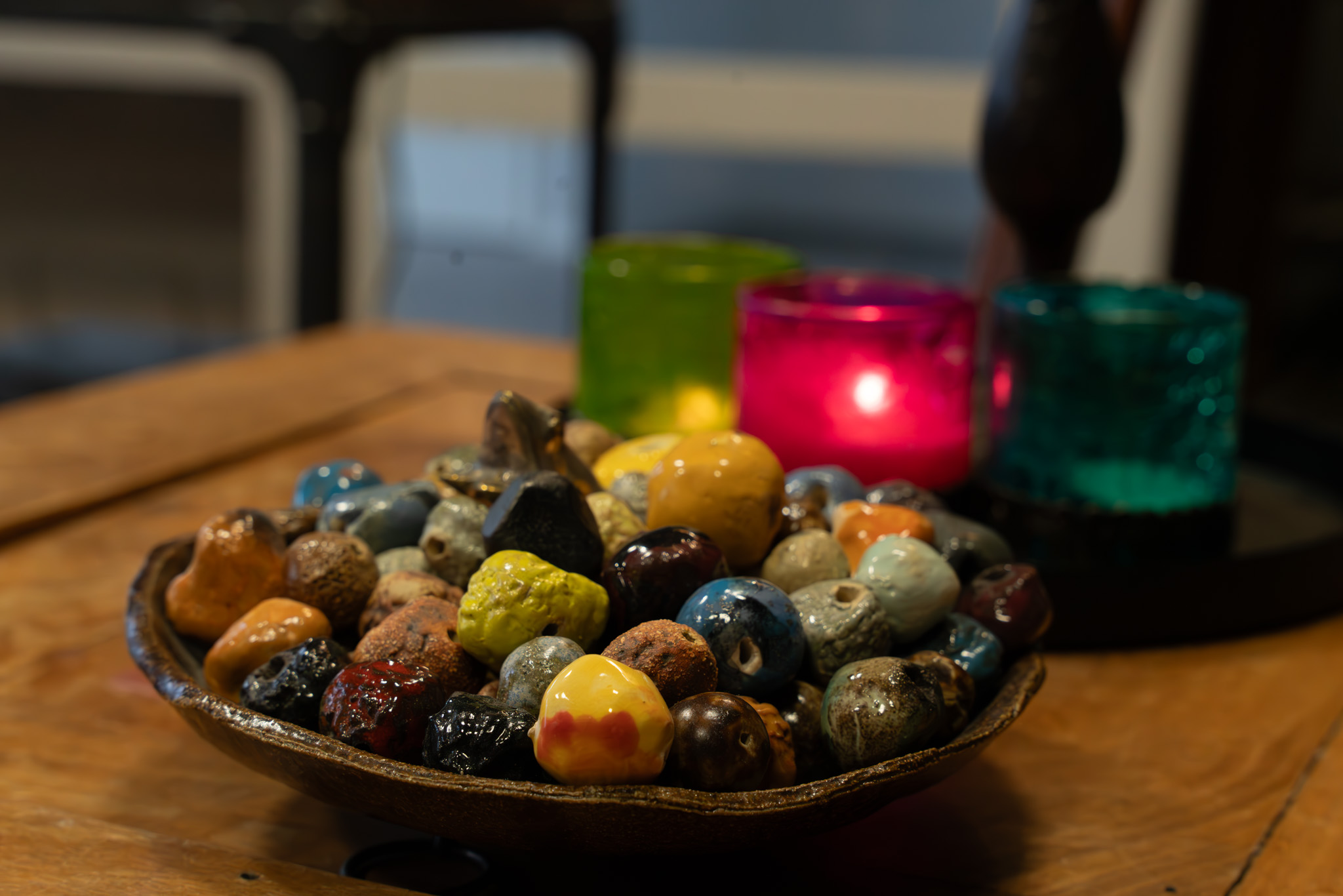 Värikkäitä kiiltäviä kiviä kulhossa, taustalla kolme värillistä, himmeästi valaistua kynttilälyhtyä.