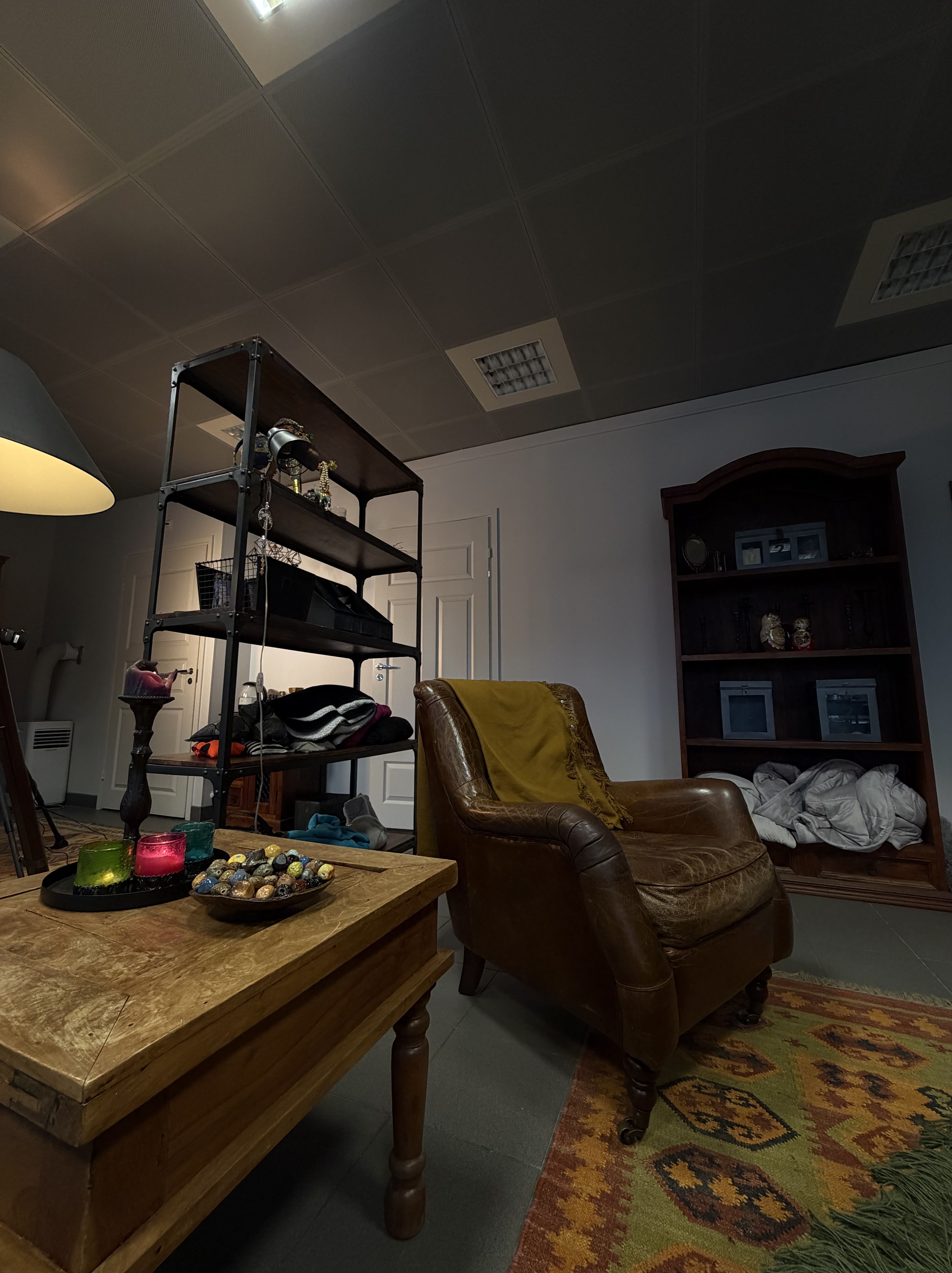 Mukava ruskea nahkainen nojatuoli keltaisella viltillä, puinen pöytä värikkäillä kynttilöillä ja kivikoristeilla, hylly täynnä vaatteita ja koriste-esineitä, sekä matto kuviollisella oranssi-vihreällä kuviolla.