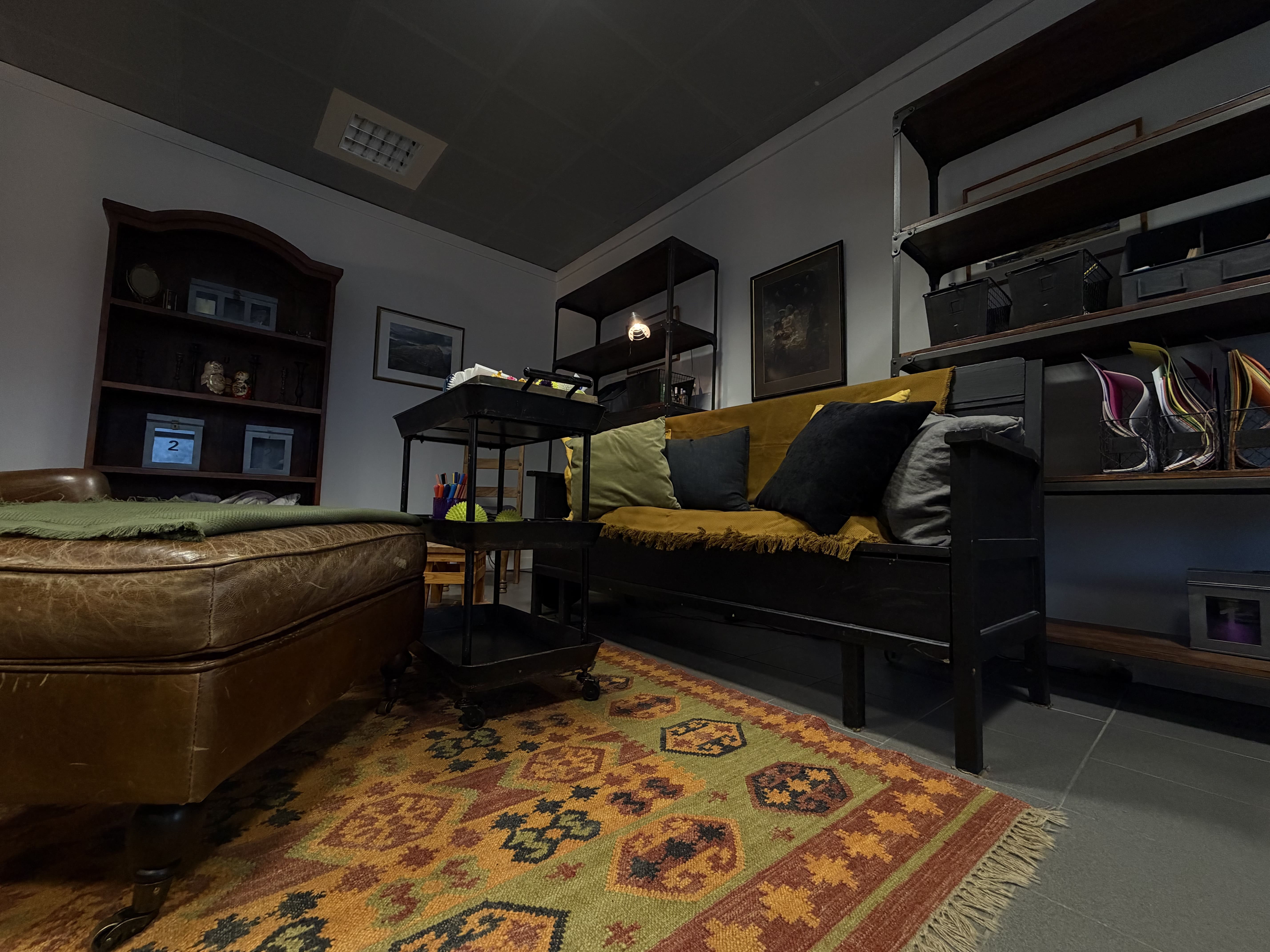 Sisätilan oleskelualue, jossa ruskea nahkasohva, musta penkki värikkäine tyynyineen ja hyllyjä tauluineen seinällä.