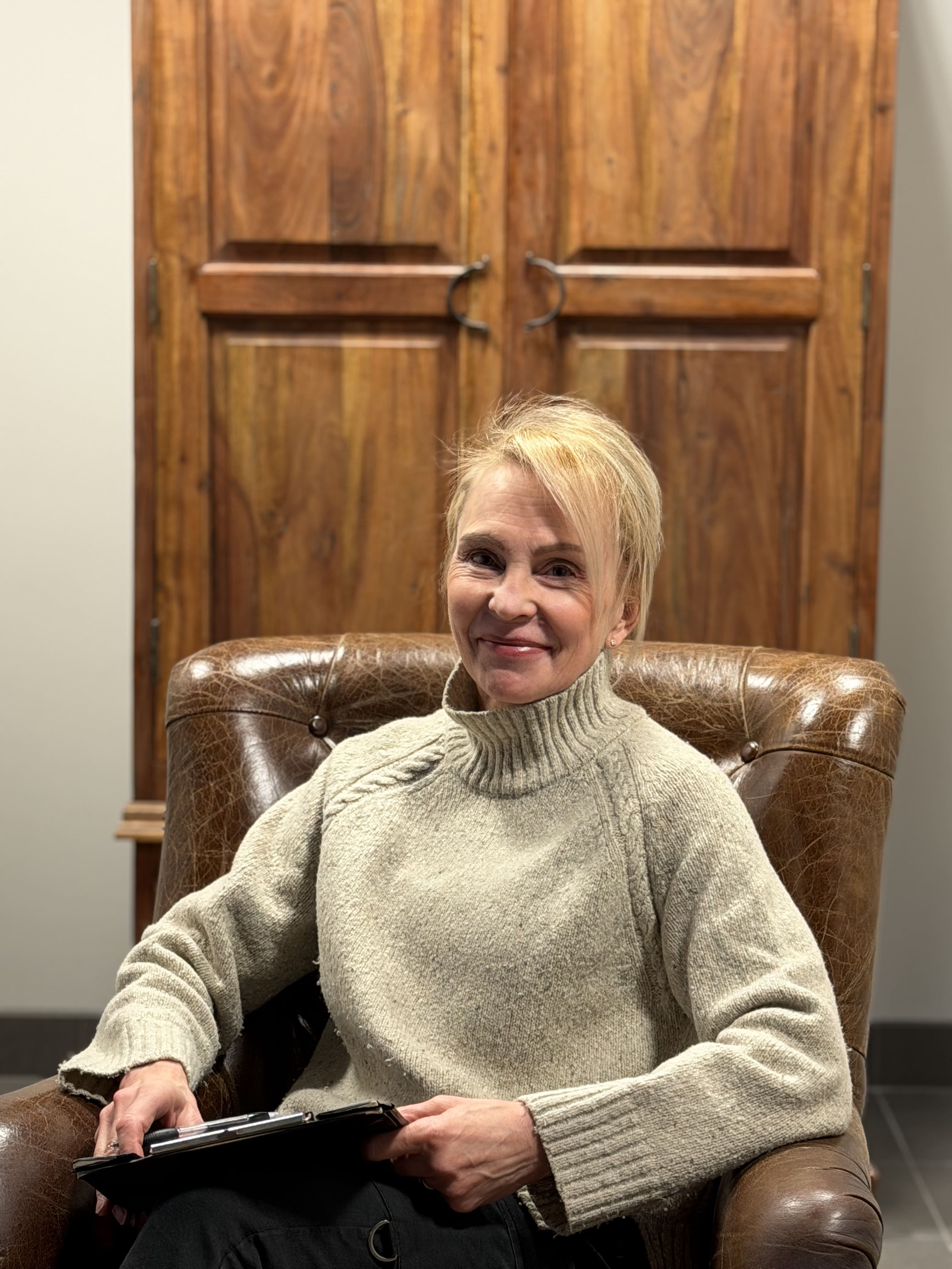 Naispuolinen henkilö istuu ruskeassa nahkatuolissa vaalean neuleen kanssa, pitäen kädessään muistikirjaa.