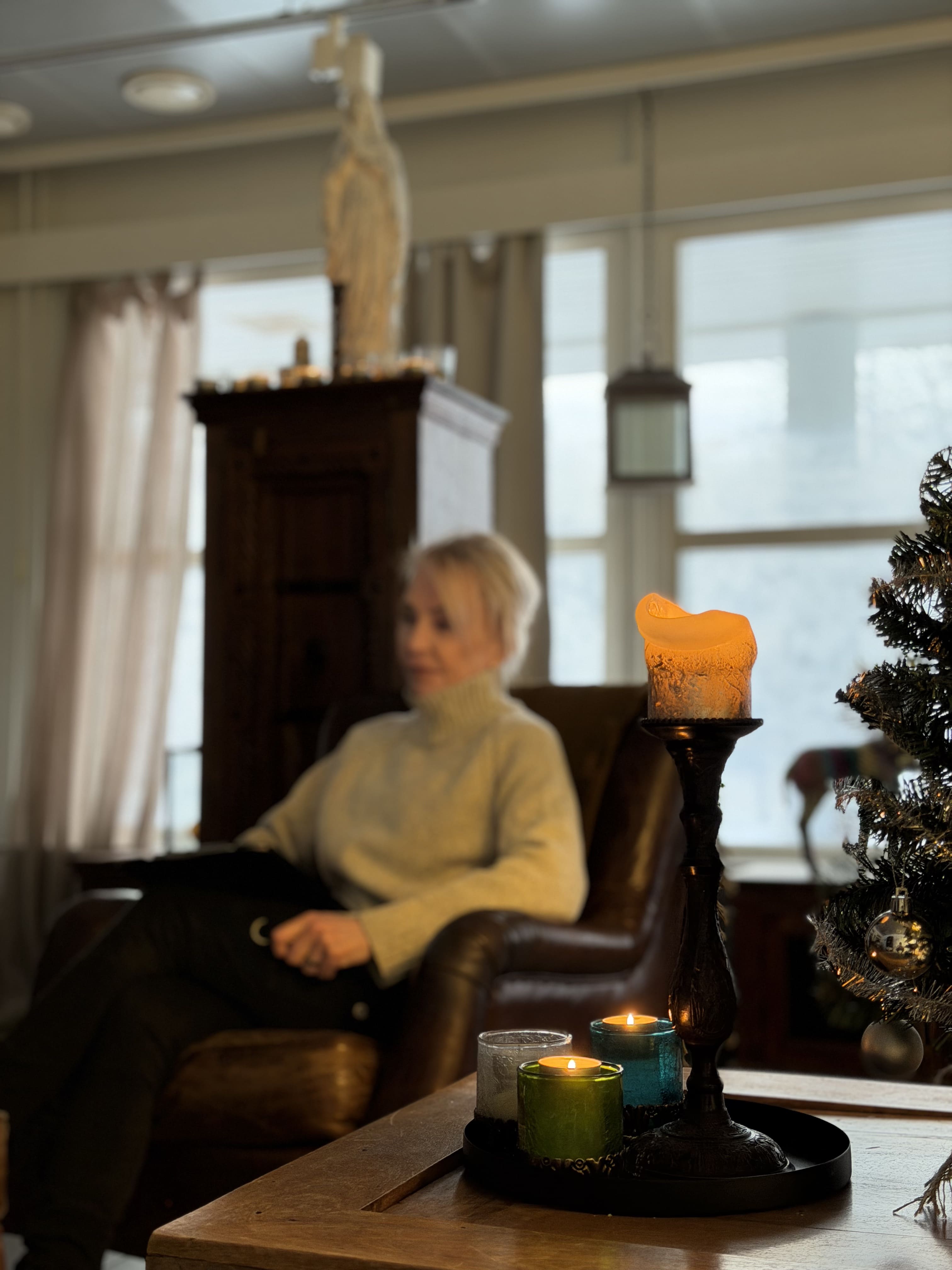 Lämmin kynttiläasetelma sohvapöydällä, taustalla nainen istuu nojatuolissa, olohuoneen sisustus sisältää joulukuusen.