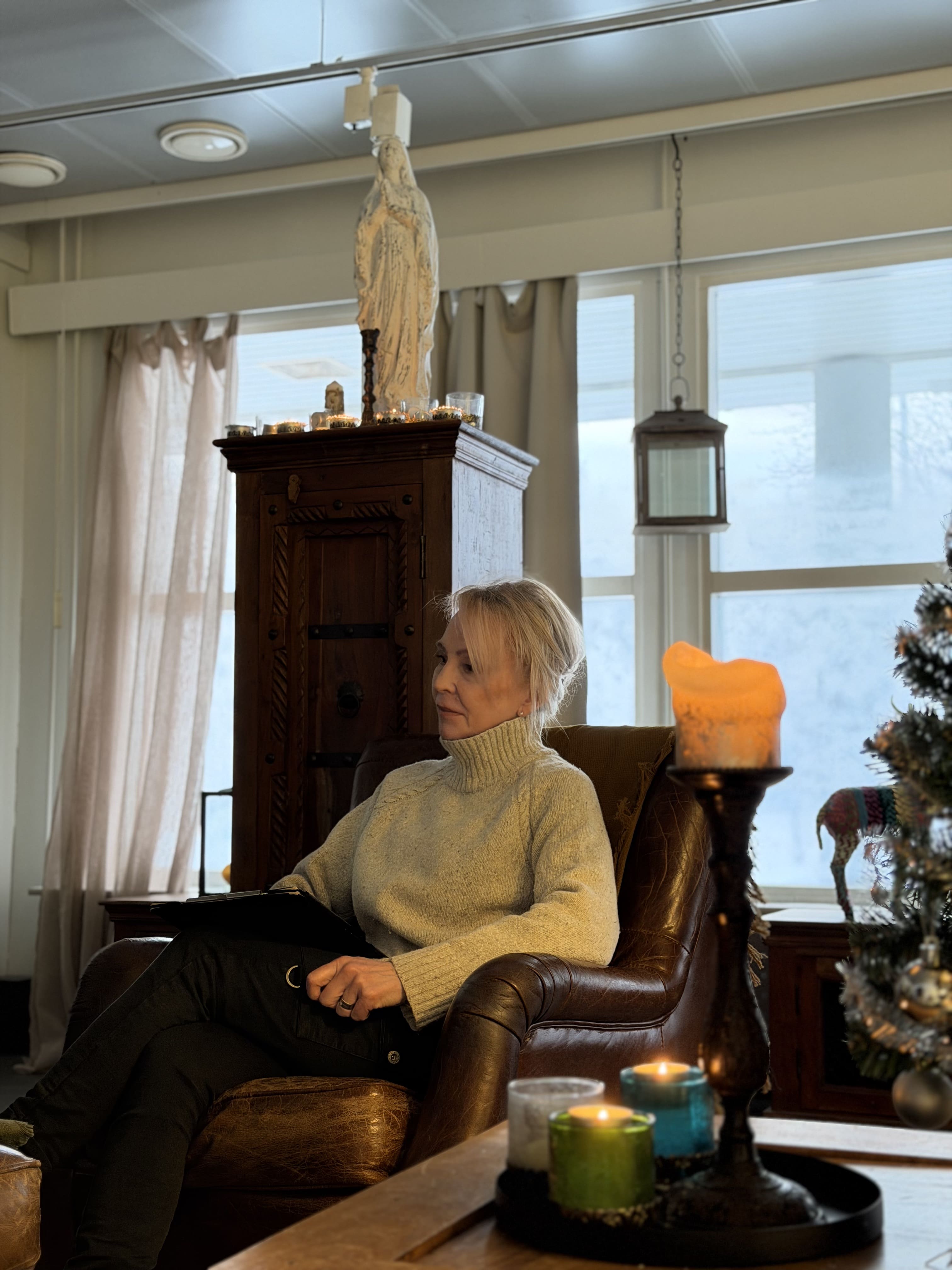 Nainen istuu nahkatuolissa ja katsoo sivulle, taustalla kynttilöitä, patsas ja jouluinen sisustus.