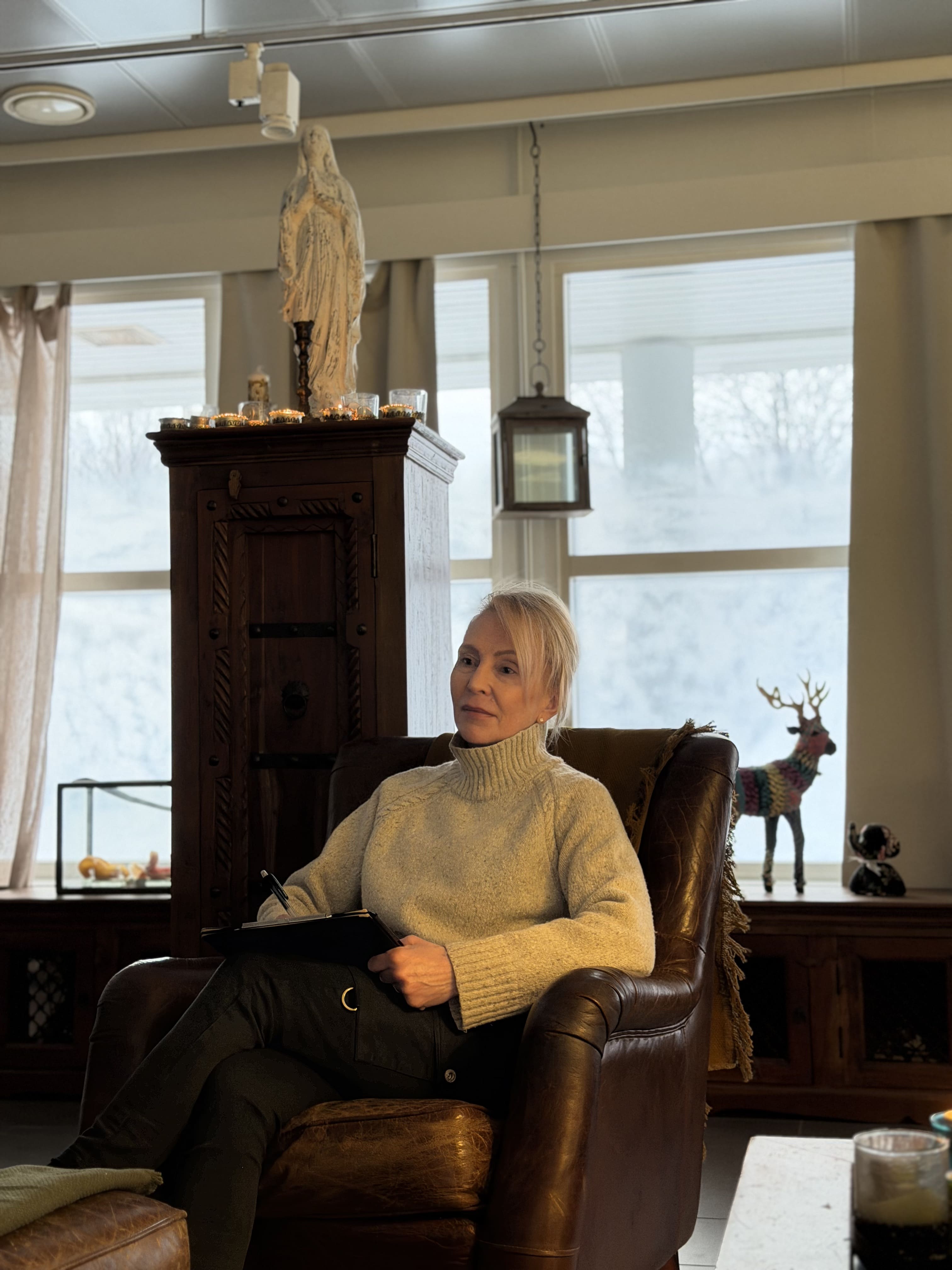 Nainen istuu ruskeassa nahkatuolissa kirjaten muistiin, takana koristeita ja suuret ikkunat.