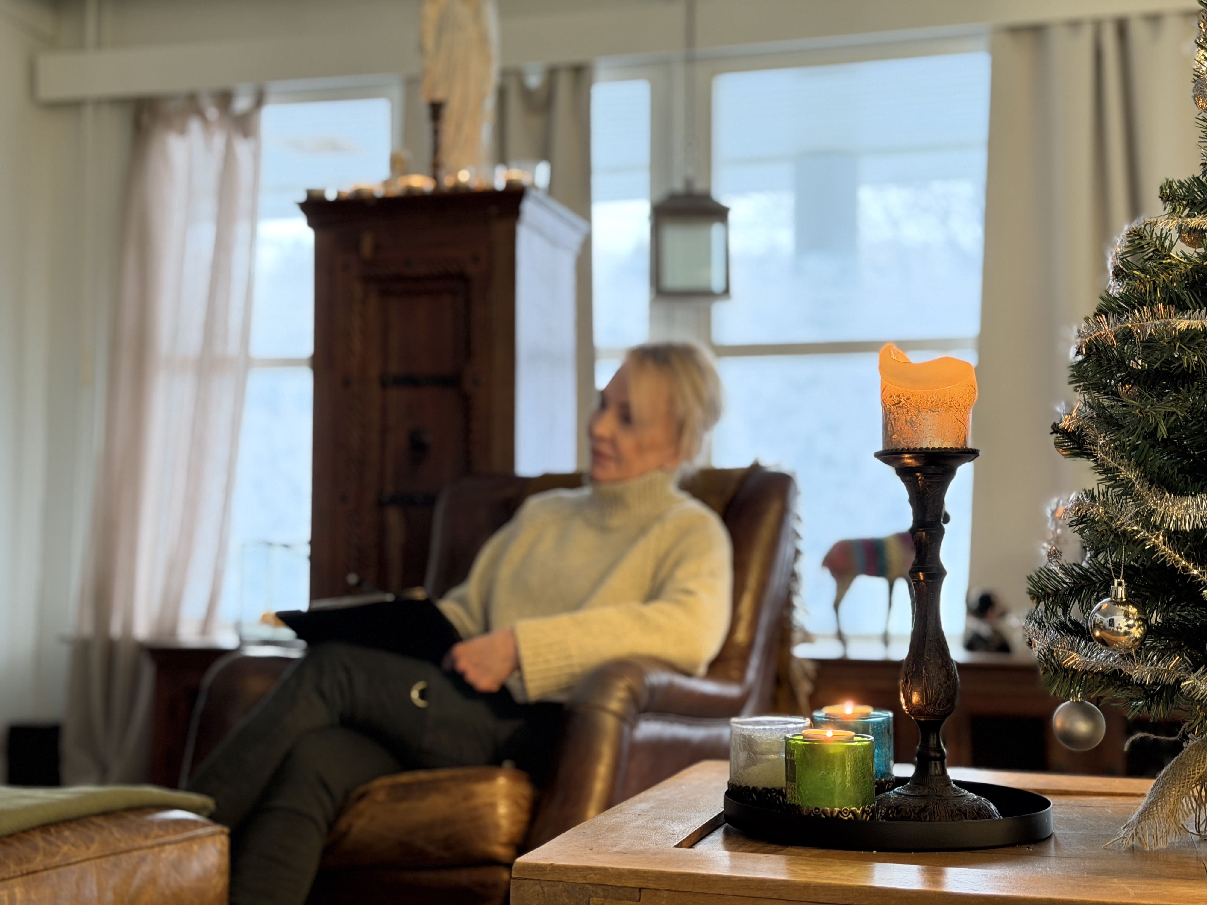 Nainen istuu nahkasohvassa ikkunan ääressä pitämässä muistikirjaa, edessä kynttilöitä ja koristeltu joulukuusi.