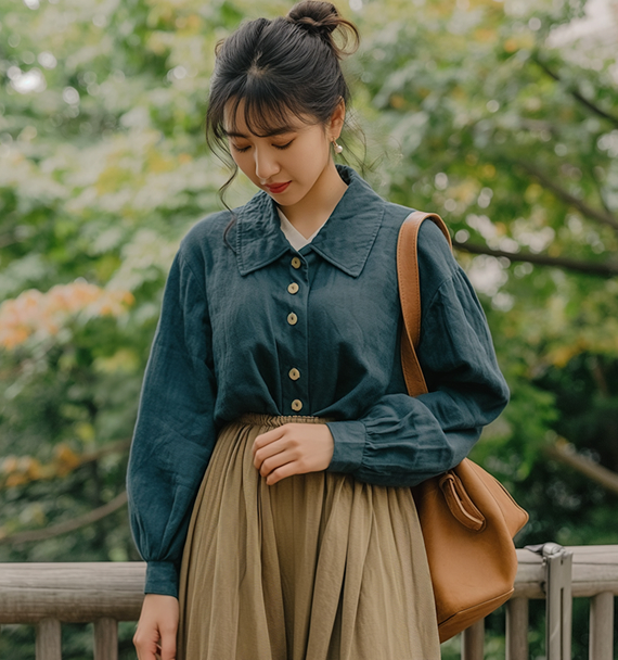 Nainen tummansinisessä napillisessa paidassa ja beigehameessa seisoo ulkona vihreän luonnon ympäröimänä kantamassa ruskeaa olkalaukkua.