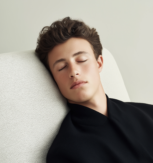 Nuori mies nojaa vaaleaan sohvaan suljetuin silmin, pukeutuneena mustaan puseroon.