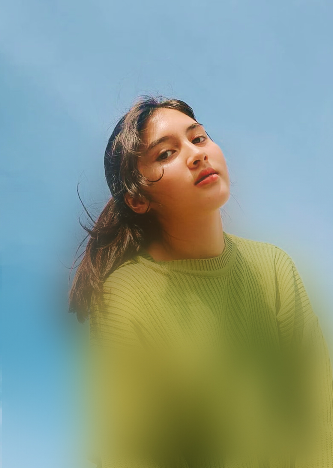 Nuori nainen vihreässä neuletakissa taivasta vasten katsomassa kameraan.