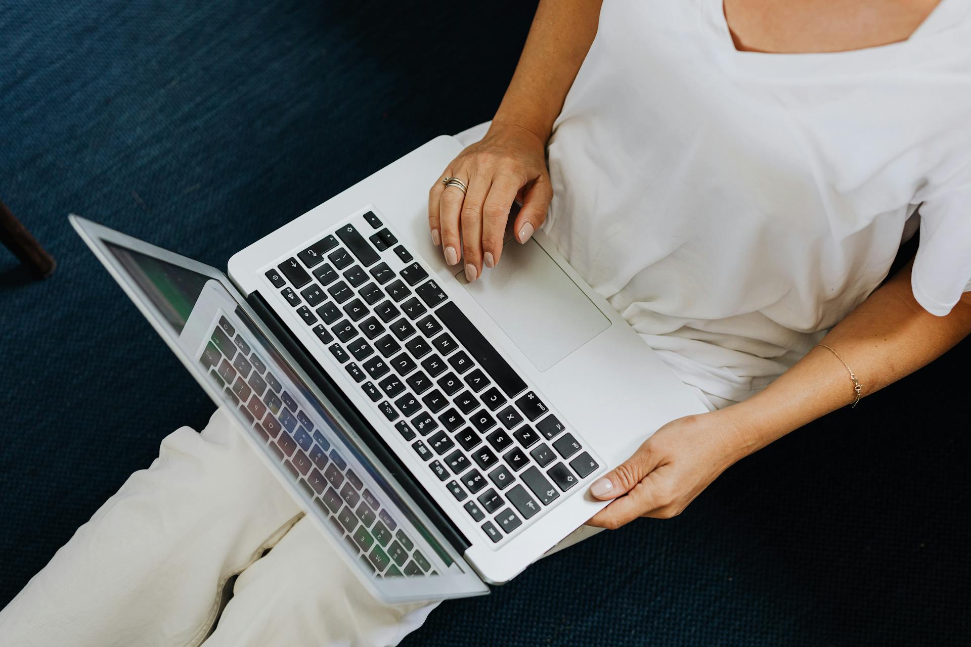 Henkilö istuu sinisellä matolla ja käyttää kannettavaa tietokonetta, jossa on hopeanhohtoinen kuori ja mustat näppäimistönäppäimet.